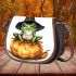 Frog wearing a black witch's hat sitting on top of a halloween pumpkin saddle bag